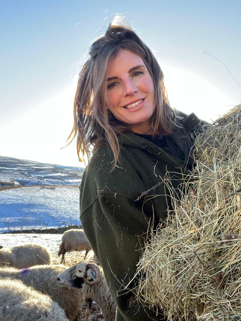 Woman smiling holding a stack of hay with sheep in the snowy fields